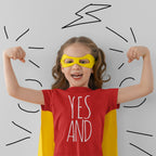 Child in superhero costume with 'YES AND' shirt flexing muscles.